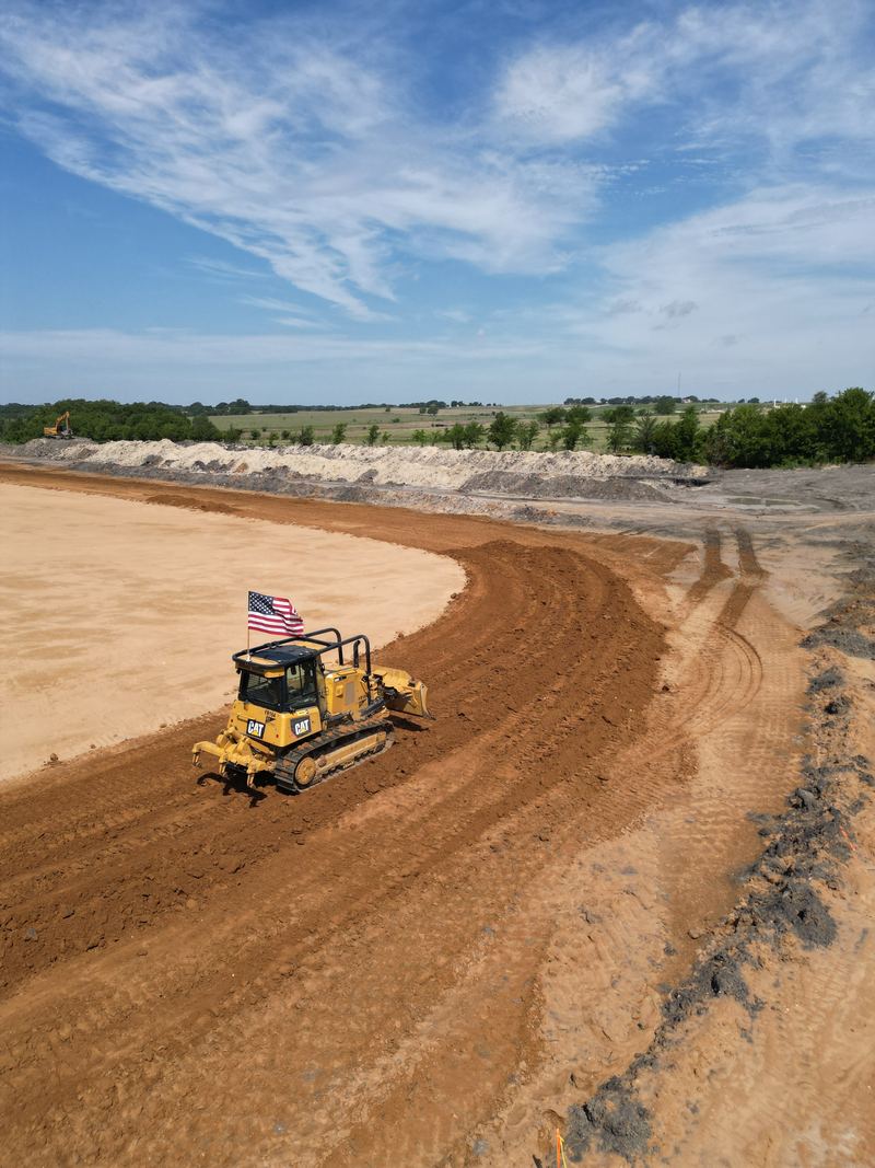 Aerial view of a dozer flying an American flag while grading a curved red-dirt pad on a school site