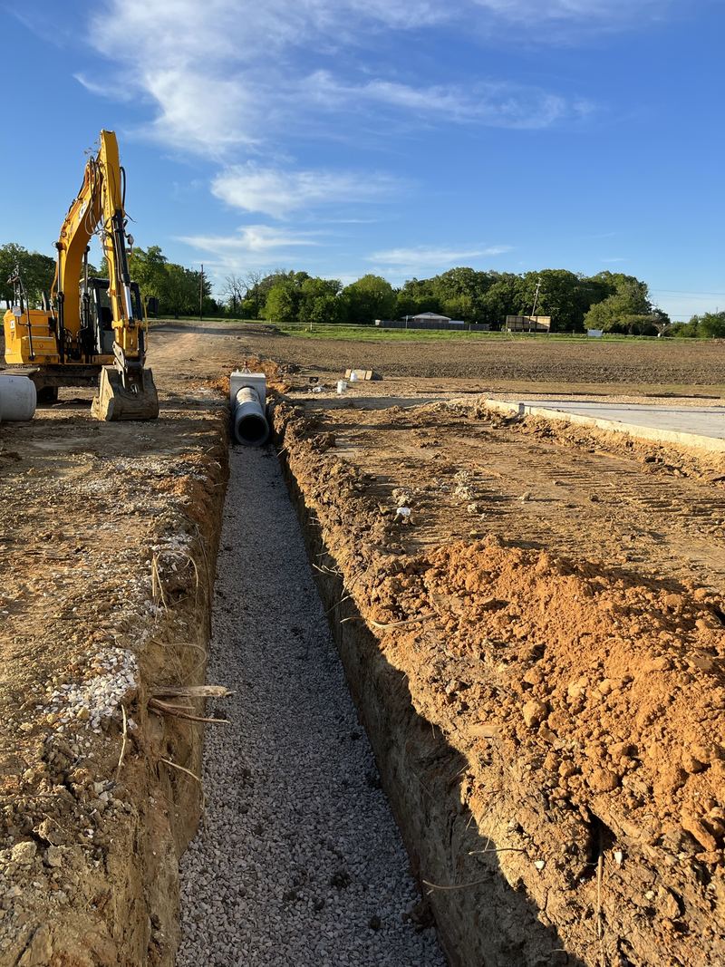 Excavator beside a long trench with a gravel bed and pipe, near Paris, TX